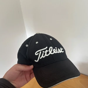 Titleist keps  - Svart golf keps från Titleist med vit broderad logga framtill. OBS Kepsen har trasigt justerbart spänne bak. Tillverkad i bomull, perfekt för golf eller andra utomhusaktiviteter.
