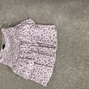 Ljuslila blommig volangkjol - Supersöt kort kjol med volanger och smock i midjan. Kjolen är ljuslila med små lila blommor över hela tyget och har en luftig, somrig känsla. Perfekt för dig som gillar romantiska och feminina plagg med mycket detaljer.