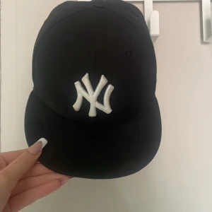Svart New Era Yankees keps - Svart keps från New Era med klassiskt New York Yankees broderat logga i vitt framtill. Kepsen har böjd skärm och broderade detaljer på sidan och bak. Perfekt för dig som gillar sportig streetstyle och vill ha en ikonisk look.