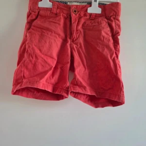 Röda shorts från Hampton Republic - Snygga röda shorts från Hampton Republic med klassisk passform och midwaist. Gjorda i mjuk bomull, har fickor både fram och bak samt knappstängning. Insidan har ett coolt prickigt mönster i blått och vitt som ger extra stilpoäng. Perfekta för varma sommardagar!