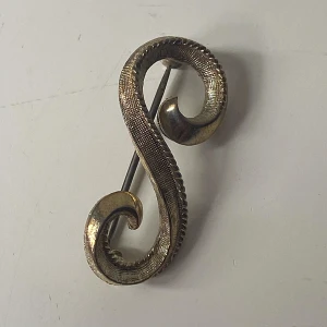 Vintage guldtonad brosch i S-form - Unik brosch i guldtonad metall med snyggt S-format, snirkligt mönster och texturerad yta. Perfekt accessoar för att ge din outfit en cool retrovibe. Cirka 4 cm lång och lätt att fästa på jacka eller väska.
