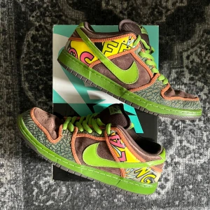 Nike SB Dunk Low De La Soul 10US - Nike SB Dunk Low De La Soul med grön och brun mocka, limegröna detaljer och snörning. Sidorna har färgglada grafiska mönster i gult, rosa och svart. Grön sula och orangea kontrastsömmar. Perfekta för dig som vill sticka ut med unika sneakers.