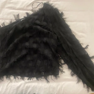 Svart fransig one shoulder-topp Gina Tricot - Säljer en svart one shoulder-topp från Gina Tricot med långa fransar över hela tyget. Toppen har en lång ärm och croppad passform som ger en cool och trendig look. Perfekt för dig som vill sticka ut med en unik detalj.