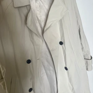 Beige trenchcoat med bälte - Stilren beige trenchcoat med klassisk krage, svarta knappar och knytskärp i midjan. Jackan har långa ärmar och axelklaffar för en extra snygg look. Perfekt för dig som vill ha en tidlös och trendig jacka till garderoben. Helt ny, aldrig använt.