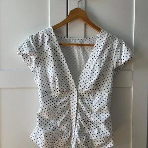 Vit prickig blus med v-ringning - Supersöt vit blus med svarta prickar, djup v-ringning och knappar framtill. Korta puffärmar och lätt rynkade detaljer vid midjan ger en feminin siluett. Perfekt för dig som gillar retrovibbar och vill sticka ut lite extra.