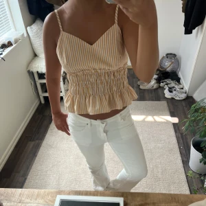 Randig croppad linnetopp med volang - Säljer en söt croppad linnetopp med smala axelband och volang nertill. Toppen har beige och vita ränder, smockad midja och v-ringad hals. Perfekt för varma dagar och ger en fräsch sommarkänsla.