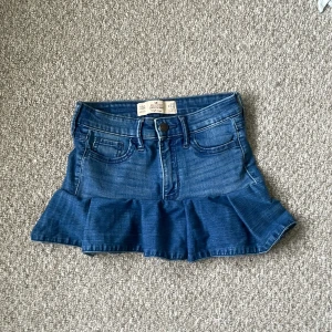 Jeanskjol med volang från Hollister - Supersöt blå jeanskjol från Hollister med volang nertill och klassiska fickor fram och bak. Hög midja och snygg denim-look som ger en cool vibe. Perfekt för dig som gillar en trendig och avslappnad stil.