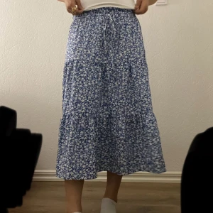 Blommig blå kjol med snörning - Supersöt blå kjol med små vita blommor och volanger. Kjolen har resår i midjan med snörning för extra bekväm passform. Perfekt flowy känsla och lätt material som passar till sneakers eller sandaler.