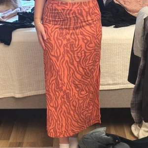 Orange zebramönstrad långkjol - Säljer en snygg långkjol (midi-ish) i orange med zebramönster i en mörkare nyans. Kjolen har en rak passform och en slits på sidan. Säljer då den är för liten i midjan för mig. Mitt midjemått är ca 75 cm. Knappt använd. Är 160 för referens. 