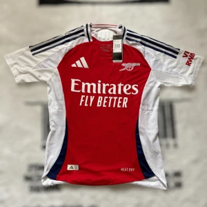 Arsenal hemmatröja Adidas Player Edition - Snygg Arsenal hemmatröja från Adidas med klassisk röd front, vita ärmar och mörkblå detaljer. Tröjan har korta ärmar, broderat klubbmärke och Adidas-logga. Tillverkad i ventilerande funktionsmaterial, perfekt för match eller träning. (Tröjan sitter lite tightare då det är player edition och inte fan version)