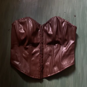 Brun skinnkorsett med markerade sömmar - Säljer en snygg brun korsett i skinnimitation med markerade sömmar och formad byst. Toppen är axelbandslös och har en stilren, edgy look som passar perfekt till jeans eller kjol.