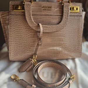 Guess ljusbrun handväska  - Guess handväska i elegant krokodilmönster med justerbar axelrem och avtagbar berlock. Väskan är i mycket fint skick, med inga eller endast minimala tecken på användning. Storlek: 30x25x21cm Material: PU och polyester Levereras med original Guess-påse. Köptca för cirka 1 600 kr (beroende på butik)