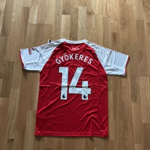 Arsenal hemmatröja Gyökeres #14 - Snygg Arsenal hemmatröja från Adidas med Gyökeres och nummer 14 på ryggen. Röd kropp med vita korta ärmar och vita detaljer, samt klubbmärke och sponsortryck på bröstet. Tillverkad i lätt och ventilerande polyester, perfekt för match eller träning.