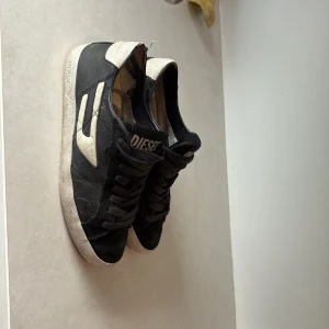 Svarta sneakers från Diesel i skinn - Snygga svarta sneakers från Diesel med vita detaljer och logga på plösen. Skorna har klassisk snörning, rund tå och är tillverkade i skinn med en vit sula. Perfekta för dig som gillar en stilren och sportig look.