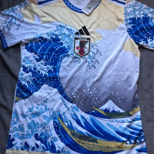 Japan Adidas fotbollströja Hokusai-våg - Japan fotbollströja från Adidas med ikoniskt Hokusai-våg motiv i blått, vitt och beige. Tröjan har korta ärmar, Japan-emblem på bröstet och klassiska Adidas-detaljer. Tillverkad i lätt och ventilerande material, perfekt för match eller träning.