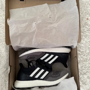 Adidas Ultraboost svarta och grå sneakers - Snygga Adidas Ultraboost sneakers i svart och grått med vita detaljer och klassiska tre ränder på sidorna. Ovandelen är i stickad syntet för skön passform och andningsförmåga. Sulan är vit och ger grym komfort. Perfekta för dig som gillar stilrena och sportiga skor.