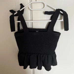 Svart linne med volang - Supersöt svart linnetopp från SHEIN med breda axelband som knyts på axlarna. Smockad överdel och volang nertill ger en trendig och feminin look. Perfekt att styla med jeans eller kjol för en cool vibe.