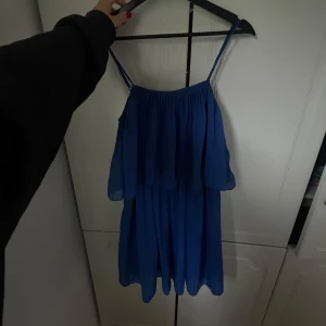 Blå axelbandsklänning från Gina Tricot - Supersöt blå kortklänning med tunna axelband och volang upptill. Klänningen är luftig och har ett flowy fall, perfekt för varma dagar. Tillverkad i ett lätt och skönt polyestertyg som ger en snygg siluett.