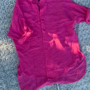 Rosa oversized skjorta i bomull - Snygg oversized skjorta i stark rosa färg med knappar framtill. Skjortan har lång ärm som kan vikas upp och är tillverkad i ett luftigt bomullstyg, perfekt för varmare dagar. Avslappnad passform och rundad nederkant ger en chill vibe.