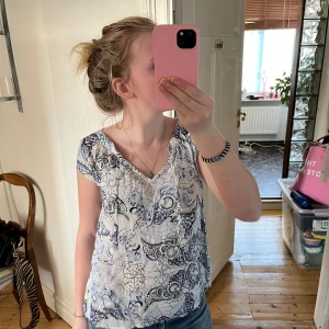 Vit och blå blommig blus med v-ringning - Säljer en luftig blus med v-ringning och korta ärmar. Toppen har ett blommigt mönster i blått och svart på vit botten, vilket ger en fräsch och somrig vibe. Perfekt till jeans eller kjol för en chill look.