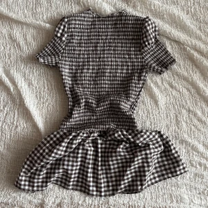 Brun och vit rutig kortklänning - Supersöt kortklänning med brunt och vitt rutigt mönster. Klänningen har puffiga korta ärmar, smockad överdel och volangig kjol. Perfekt för dig som gillar en lekfull och trendig stil. Materialet känns lätt och andas, troligtvis bomull.