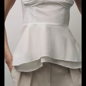 Vit peplum linnetopp med knytband - Supersöt vit linnetopp med peplum och tunna axelband som knyts i ryggen. Toppen har en figurnära passform upptill och volangig nederdel i dubbla lager. Perfekt för varma dagar och ger en romantisk vibe till din outfit.