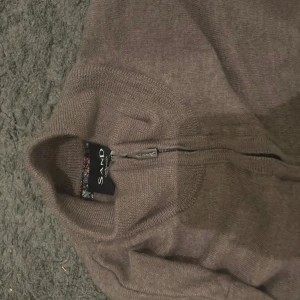 Brun stickad half zip-tröja Sand - Brun stickad tröja från Sand med half zip och hög krage. Tröjan har lång ärm och ribbade muddar vid ärmslut och nederkant. Perfekt för lager-på-lager och chill höst- eller vinterstil.