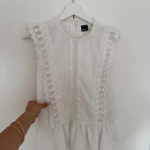 Vit spetsblus från Gina Tricot - Supersöt vit blus från Gina Tricot med romantiska spetsdetaljer längs axlar och framtill. Blusen är ärmlös, har rund halsringning och volang nertill. Perfekt för dig som gillar en feminin och trendig stil.