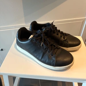 Svarta sneakers i skinn med vit sula - Stilrena svarta sneakers i skinn med klassisk vit sula och svarta snören. Låg modell med rund tå och diskreta perforeringar på sidorna. Perfekta för dig som gillar en clean och tidlös look.