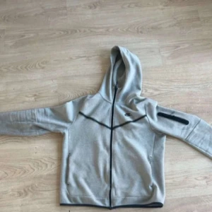 Grå Nike hoodiejacka med dragkedja - Snygg grå hoodiejacka från Nike med hel dragkedja och svarta detaljer. Jackan har huva, fickor på sidorna och en extra ficka med dragkedja på ärmen. Perfekt för chill eller träning, stilren och enkel att matcha. Har lite prickiga hål, dock inget som påverkar stilen. Bilder kan skickad önskemål.