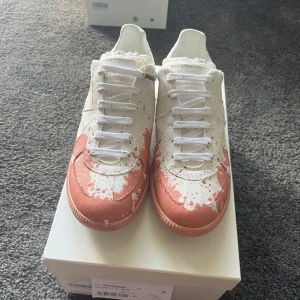 Maison Margiela Gats paint splatter - Maison Margiela Replica sneakers i vitt med unika röda färgstänk. Skorna är helt nya och aldrig använda, kommer med alla tillbehör! Perfekta skor att ha nu i sommar! Skorna är självklart autentiska och har kvitto! Storleken är 40 men passar lite större! Hör av er vid intresse!