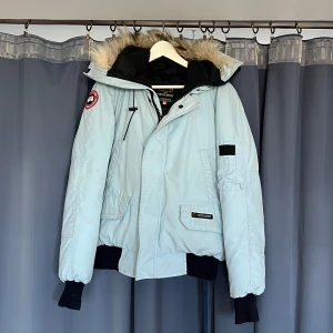 Ljusblå Canada Goose dunjacka med päls - Säljer en ljusblå dunjacka från Canada Goose med ikonisk patch på ärmen. Jackan har huva med päls, svarta muddar och dragkedja framtill. Fodrad insida och flera fickor gör den både snygg och praktisk. Perfekt för kalla vinterdagar! Nypris ca 9-12000kr