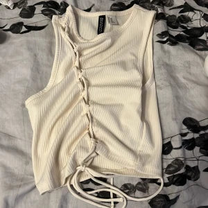 Vit ribbad linnetopp från H&M Divided - Säljer en vit ribbad linnetopp från H&M Divided i storlek XXS. Toppen är ärmlös och har snörning längs ena sidan som ger en snygg och trendig look. Perfekt för dig som gillar detaljer och vill ha något som sticker ut lite extra.