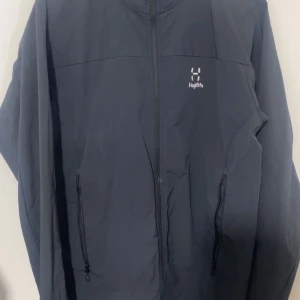 Svart vindjacka från Haglöfs - Snygg svart vindjacka från Haglöfs med dragkedja framtill och två praktiska fickor med dragkedja. Jackan har en stilren design och är perfekt för blåsiga dagar. Liten logga på bröstet ger en sportig touch.