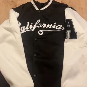 Svartvit bomberjacka från Romwe - Snygg svart och vit bomberjacka från Romwe med broderad text 'California' på bröstet och stort A på ärmen. Jackan har tryckknappar framtill och klassisk college-stil. Perfekt för dig som gillar streetwear och vill sticka ut.