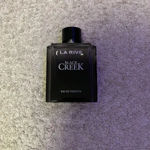 La Rive Black Creek Eau de Toilette - La Rive Black Creek Eau de Toilette i en stilren, svart glasflaska med silverdetaljer. Flaskan är fyrkantig med rund svart kork och har en modern, maskulin look. Perfekt för dig som gillar eleganta och klassiska dofter. Helt ny den är aldrig använd. 
