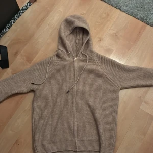 Beige stickad hoodie med dragkedja - Mysig beige stickad hoodie med dragkedja framtill och huva med snörning. Tröjan har ribbad struktur och långa ärmar, perfekt för lager-på-lager under kyliga dagar. Enkel och stilren design som funkar till det mesta.