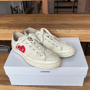 Comme des Garçons Play x Converse Chuck Taylor All Star 70 OX låga sneakers - Snygga vita Converse Chuck Taylor All Star sneakers i samarbete med Comme des Garçons Play. Skorna har det ikoniska röda hjärta-motivet på sidan, svarta detaljer baktill och klassisk snörning. Tillverkade i canvas för en clean och stilren look. Synligt använda, men i bra skick.