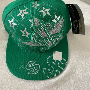 Grön keps med silvriga detaljer från Leader - Grön keps med stora silvriga dollar- och stjärnmotiv, glittriga detaljer och broderier. Märkt med Leader Sports Cap på insidan. Skärmen är platt och kepsen har en cool streetstil som sticker ut.