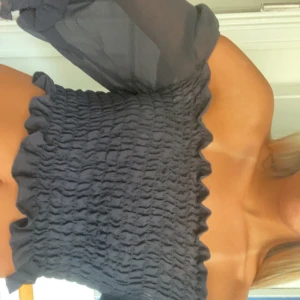 Svart offshoulder topp med puffärm - Trendig svart offshoulder topp med smockad kropp och en lång puffärm i tunnare tyg. Toppen är croppad och har en vågig kant upptill och nedtill. Perfekt för dig som vill sticka ut med en cool och modern look.