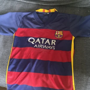 FC Barcelona Messi 10 matchtröja - Klassisk FC Barcelona fotbollströja med Messi 10-tryck på ryggen. Tröjan har breda röda och marinblå ränder, gula detaljer och klubbmärke broderat på bröstet. Kortärmad modell i lätt och ventilerande material, perfekt för fotbollsplanen eller som supporterplagg.