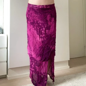 Långkjol i lila mönster - Säljer en långkjol i starkt lila och rosa toner med abstrakt mönster. Kjolen har en lätt transparent känsla, oregelbunden nederkant och slits framtill. Materialet är tunt och flowy, perfekt för dig som vill sticka ut med färg och cool struktur.