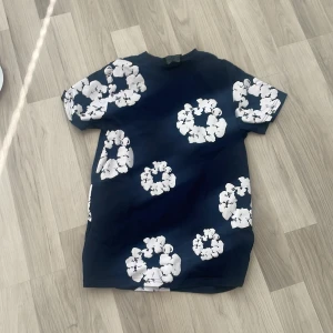 T-shirt från Denim Tears - Snygg mörkblå t-shirt från Denim Tears med stora vita blommiga tryck över hela plagget. Klassisk passform med rund hals och korta ärmar. Perfekt för dig som vill sticka ut med en unik och trendig look, mest för streetwear stilen eller mer casual. Tröjan är i storlek M och är inköpt för ungefär 2 månader sen, tröjan hängde bara kvar i garderoben och har aldrig använts sen inköp. Priset just nu är 250 sek, diskuterbart och ej hugget i sten. Hör av er vid minsta fundering! 