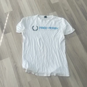 Vit Fred Perry t-shirt XL - Vit t-shirt från Fred Perry i storlek XL med blå logga och tryck framtill. Klassisk passform med korta ärmar och rund hals. Tillverkad i mjuk bomull, perfekt för en clean och enkel stil. Det finns en liten defekt ovanför loggan, men de är endast ett litet hål som inte är märkbart när tröjan bärs. Fick den av en släkting och passar inte mig alls, så väljer att sälja istället. Använd ett tiotal gånger, därför priset. Hör av er vid minsta fundering! 