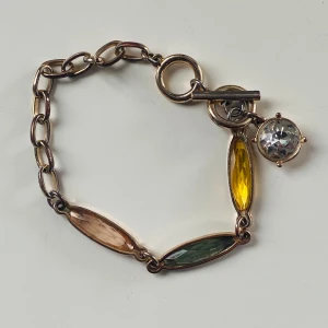 Färgglatt armband med stenar - Unikt armband med kedja i guldfärgad metall och tre avlånga, fasettslipade stenar i gult, grönt och ljusbrunt. Armbandet har en rund, klar sten som charm och stängs med en snygg T-lås. Perfekt för dig som vill ha något färgstarkt och personligt.