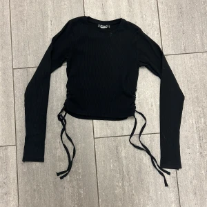 Svart ribbad topp med snörning FB Sister - Svart långärmad topp från FB Sister i ribbad bomullsmix. Croppad modell med cool snörningsdetalj i sidorna som kan justeras för perfekt passform. Perfekt till jeans eller kjol för en trendig look.