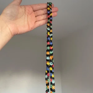 Färgglatt pärlhalsband med blommor - Unikt handgjort halsband med små pärlor i svart, rött, blått, grönt, gult, orange och vitt. Pärlorna är formade till blommor längs hela halsbandet och ger en lekfull och färgstark look. Perfekt accessoar för att lyfta vilken outfit som helst.
