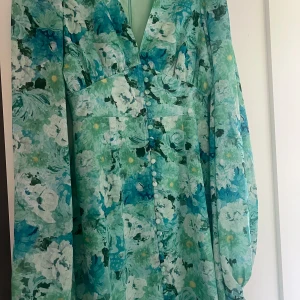 Blommig långklänning i grönt och blått - Superfin långklänning med blommigt mönster i gröna, blå och vita toner. Klänningen har långa ärmar, v-ringning och knappar framtill. Luftigt material som passar perfekt för vår och sommar. 