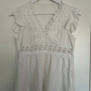 Vit blus med spets och broderi - Superfin vit blus med v-ringning, spetsdetaljer och broderade hålmönster. Blusen har volangärmar och ett dekorativt band i midjan. Perfekt för vår och sommar när du vill ha en romantisk och luftig look.