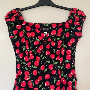 Körsbärsmönstrad top Collectif XL - Supersöt top från Collectif med röda körsbär. Den har två dekorativa knappar framtill och dragkedja i sidan för bästa  passaform. Perfekt för dig som gillar retrostil och färgglada prints.